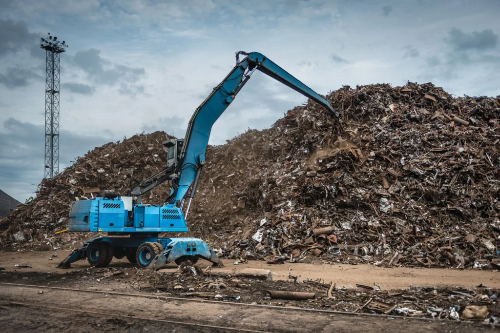 Blue excavator sorting a large pile of scrap metal at a recycling yard.