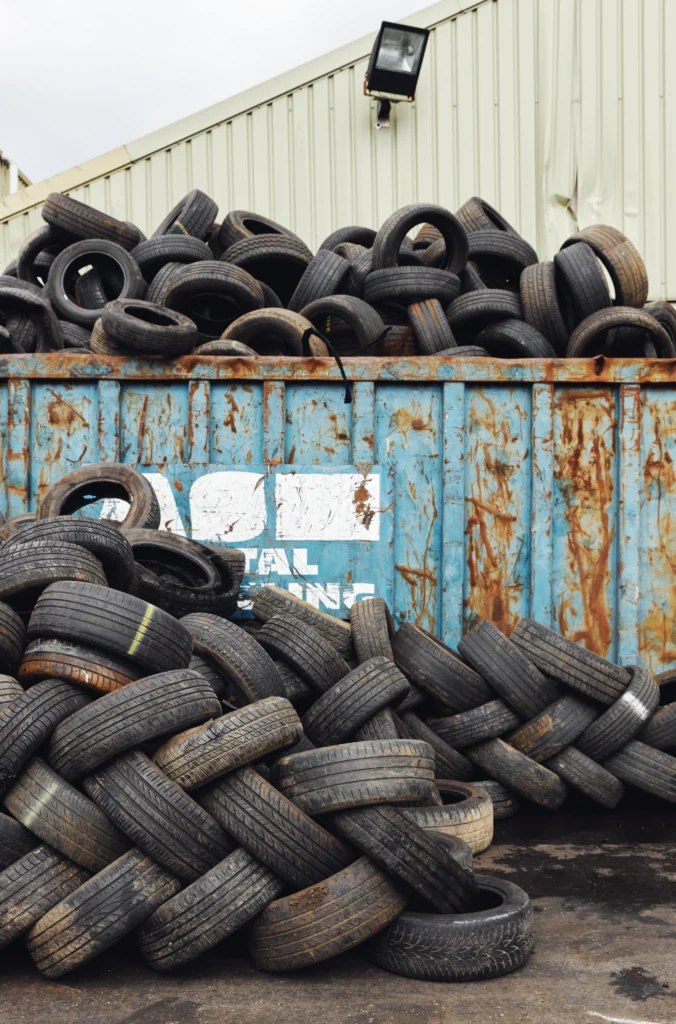 Used car tyres piled in and around a rusty blue skip at a recycling site.